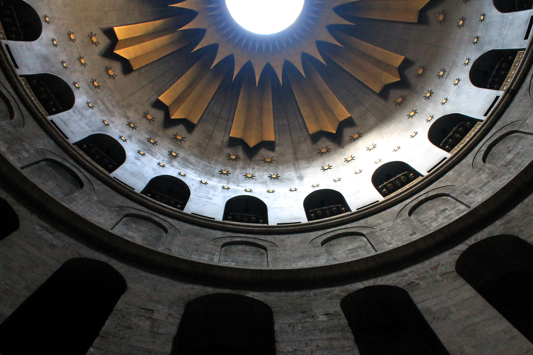 The Church of the Holy Sepulchre in Jerusalem, Israel. The image focuses on the dome of the church. The oculus is in the centre and surrounding it are rays of gold like the sun. Beneath the rays are gold stars on grey tile. There are arched doorways in the tile and also in the stone below.