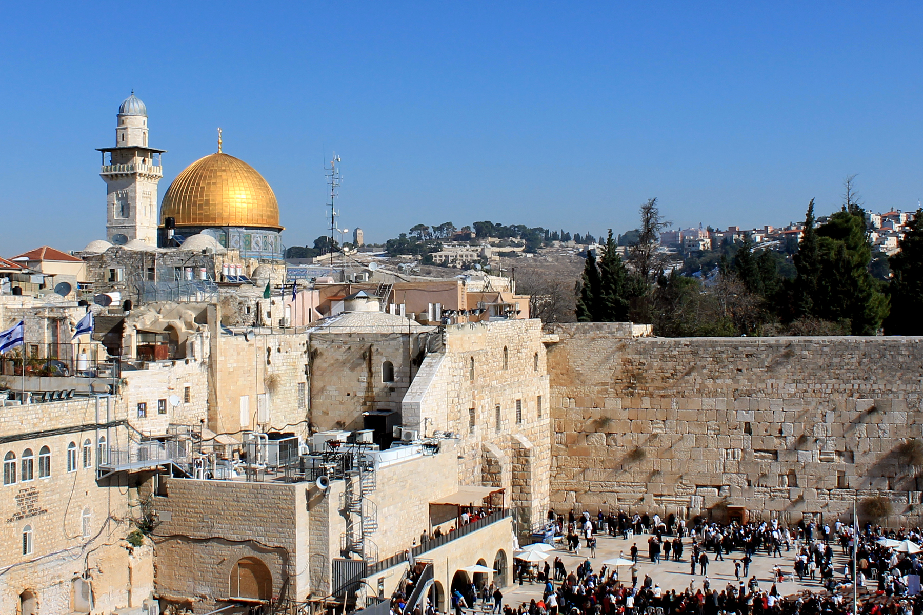 The Western Wall in Jerusalem, Israel. The image focuses on the Western Wall. The photograph has been take from a high angle and many people are waiting below to visit the wall. In the background are the hills of Jerusalem with houses and trees. In the left of the image is the Gold Dome of the Rock and a minaret tower.