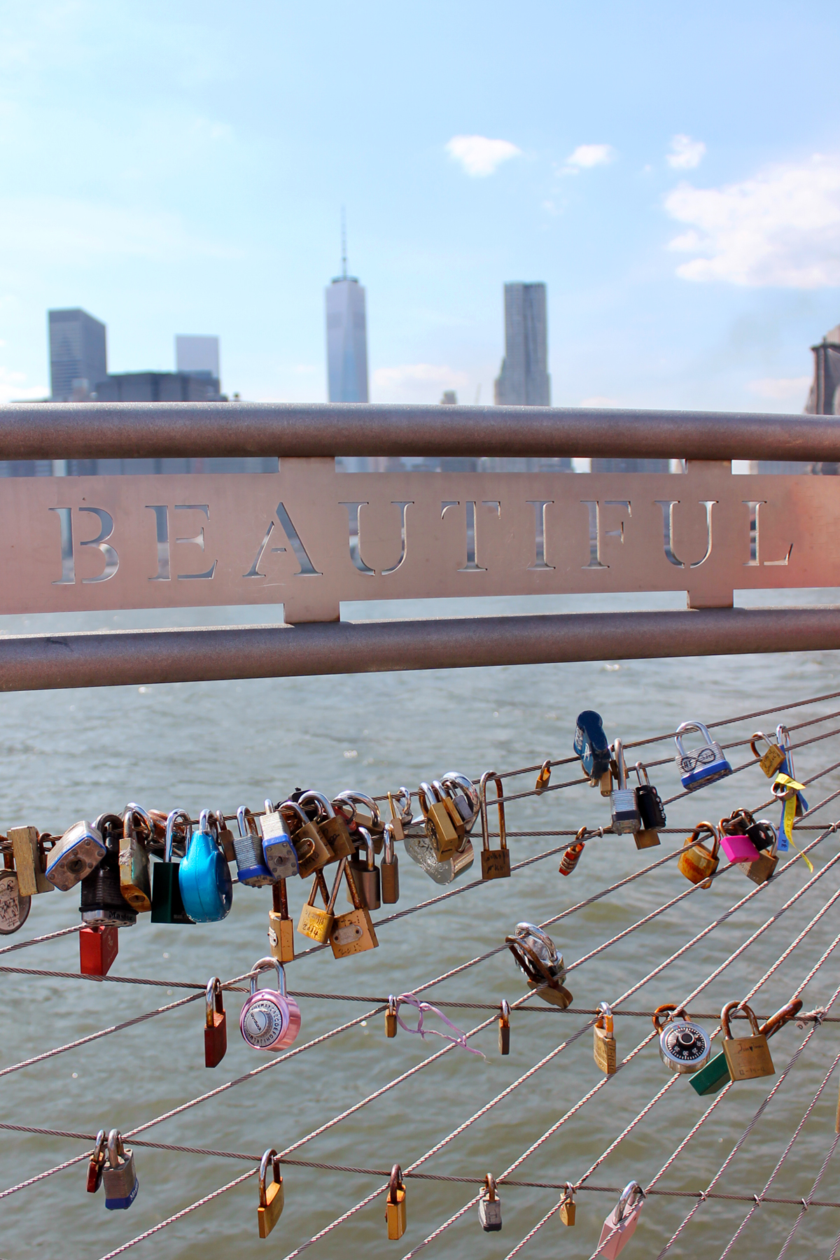 At Pier 1 in Brooklyn, New York. The New York City skyline is visible in the background. In the foreground is a closeup of a metal fence along the pier where people have attached locks. The fence reads Beautiful and the Hudson River is visible through the gaps.