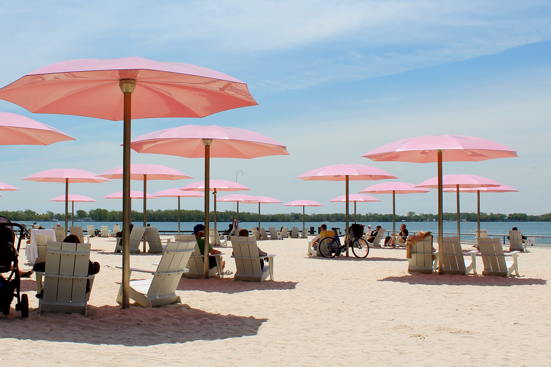 Sugar Beach in Toronto, Canada on a bright, sunny day. The sky is blue with some wispy clouds. The beach has a sea of pink umbrellas and white adirondack chairs on the white sand. Some of the chairs are occupied by people who are facing Lake Ontario.