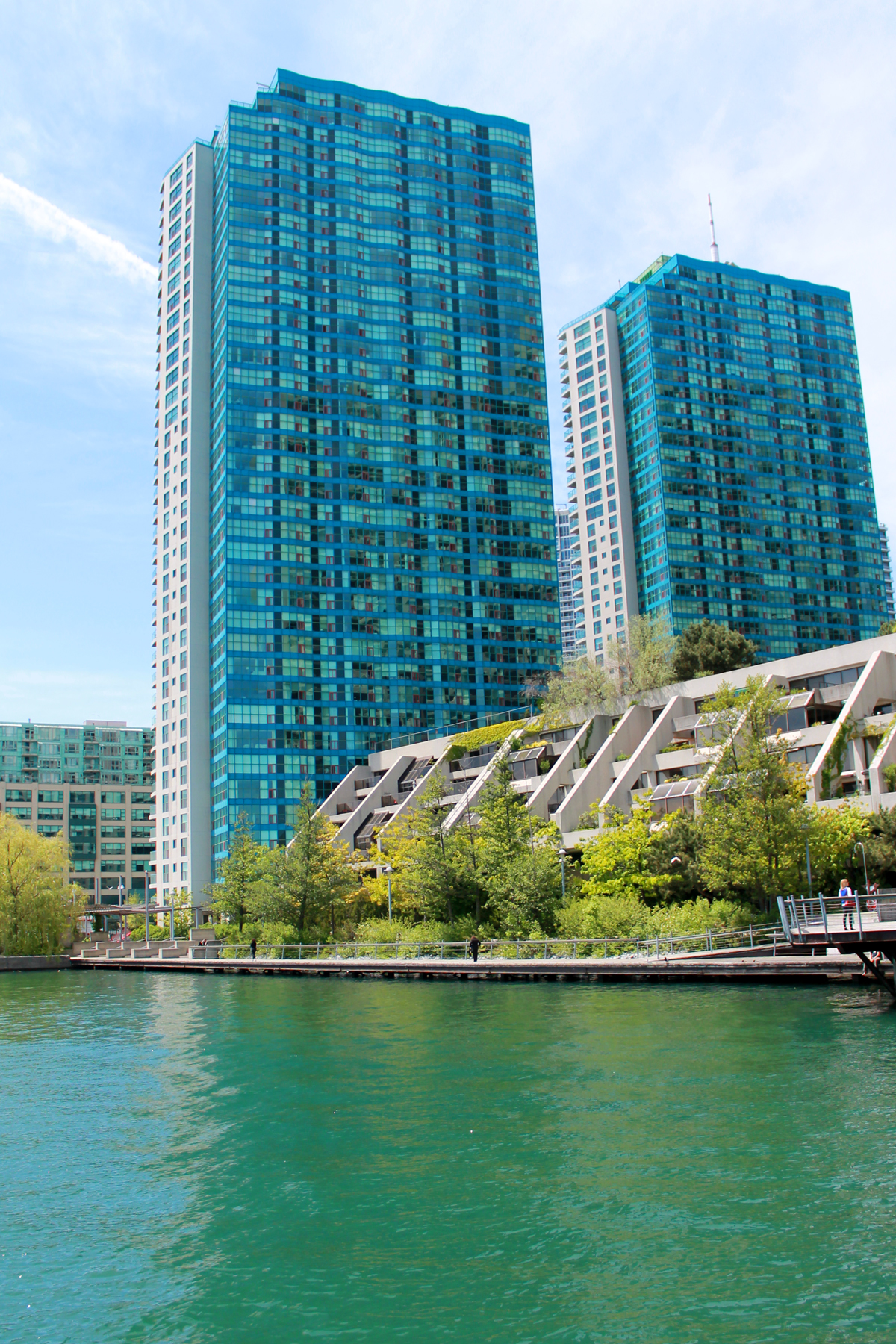 Harbour Square in Toronto, Canada on a bright, sunny day. The sky is blue with some wispy clouds. In the middle of the image are two large apartment buildings with blue glass windows of the York Quai apartment complex. In front is a concrete structure with trees and grass and the sidewalk bordering Lake Ontario.