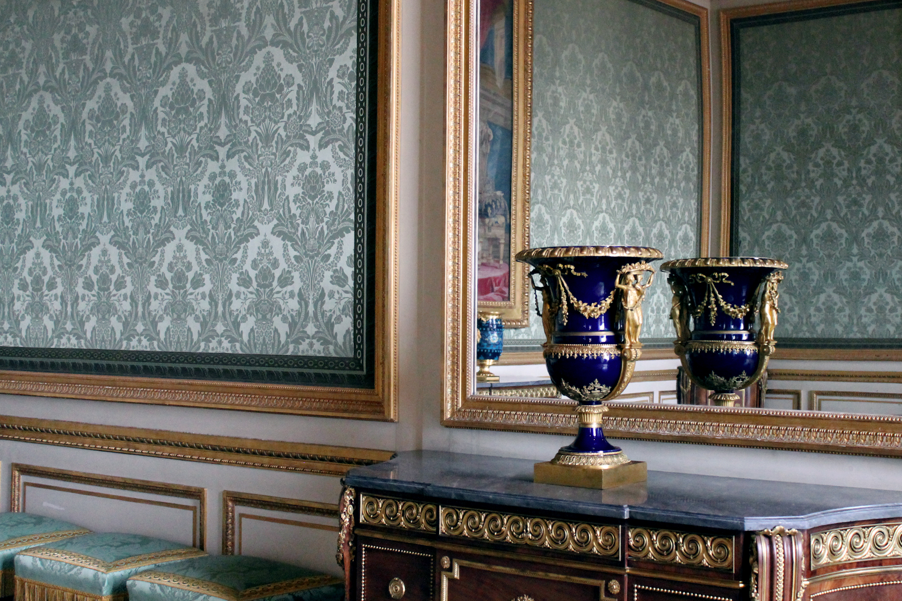 The Château de Versailles in Versailles, France. This image was taken inside a room with green patterned wallpaper and focuses on a gold and blue vase sitting on a wooden sideboard with a black top and gold detailing. Behind the vase is a large mirror with a gold frame.