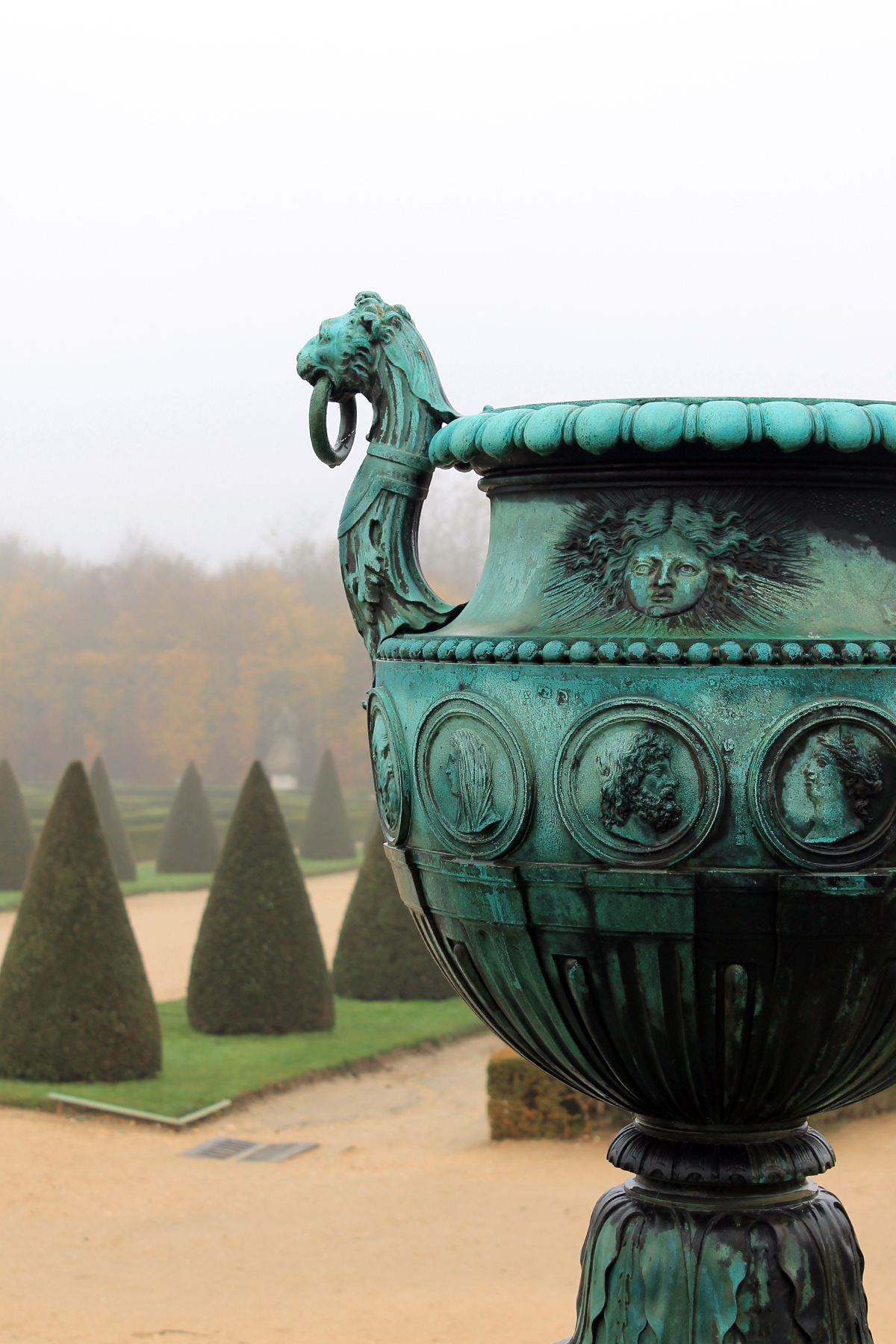 The garden of Versailles in Versailles, France on a foggy day. The image focuses on a copper vase in the garden that has turned green. The vase has decorative faces and a lion’s head. In the background are paths and narrow strips of green grass with perfectly conical trees.